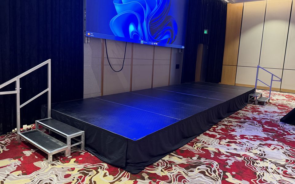 Black-skirted stage with handrailed steps in front of an LED screen in a function room