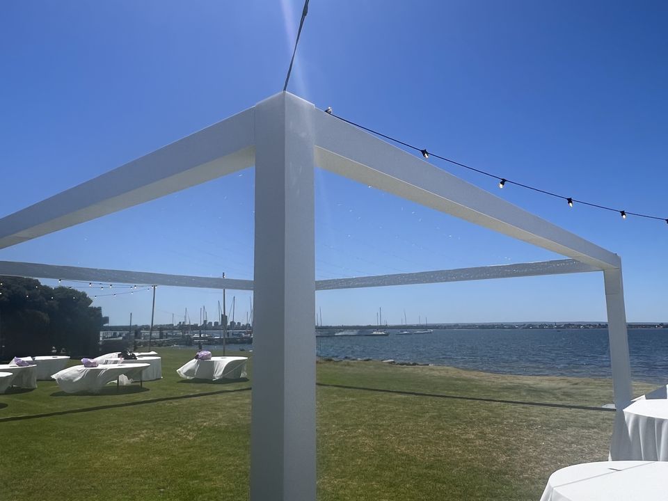 White box truss frame on a waterside lawn, with festoon lights strung from it